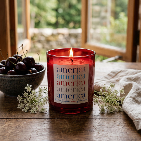 America Candle burning on a rustic farmhouse table by an open summer window | Noteology