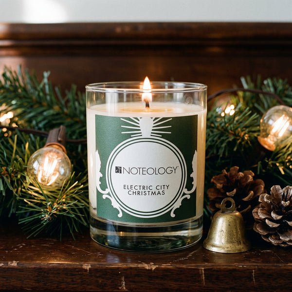 Electric City Christmas Candle burning on a dark wooden mantel draped with pine garland | Noteology