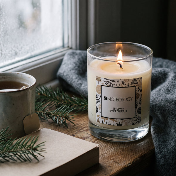 Snowy Evergreen Candle burning on a worn wooden window seat with a folded grey wool blanket beside a frosty window | Noteology