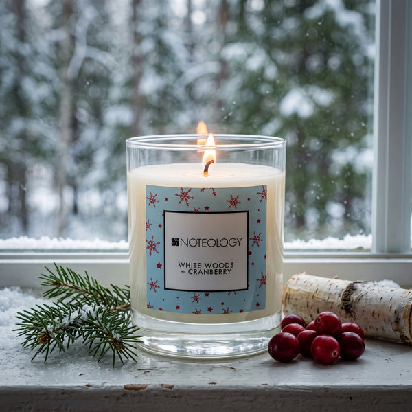 White Woods + Cranberry Candle burning on a snowy windowsill, with a blurred snowy evergreen forest outside in cool daylight | Noteology